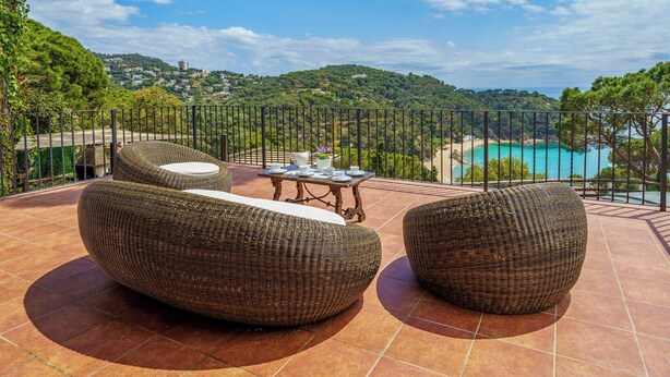 Een terras met rieten meubilair, een tafel met kopjes en schotels, en uitzicht op een kustlandschap.