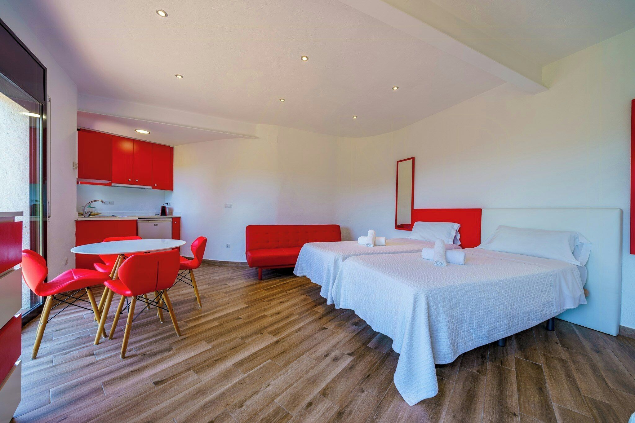 Une chambre d’hôtel avec une kitchenette, un coin repas avec des chaises rouges et un lit avec du linge de lit blanc.