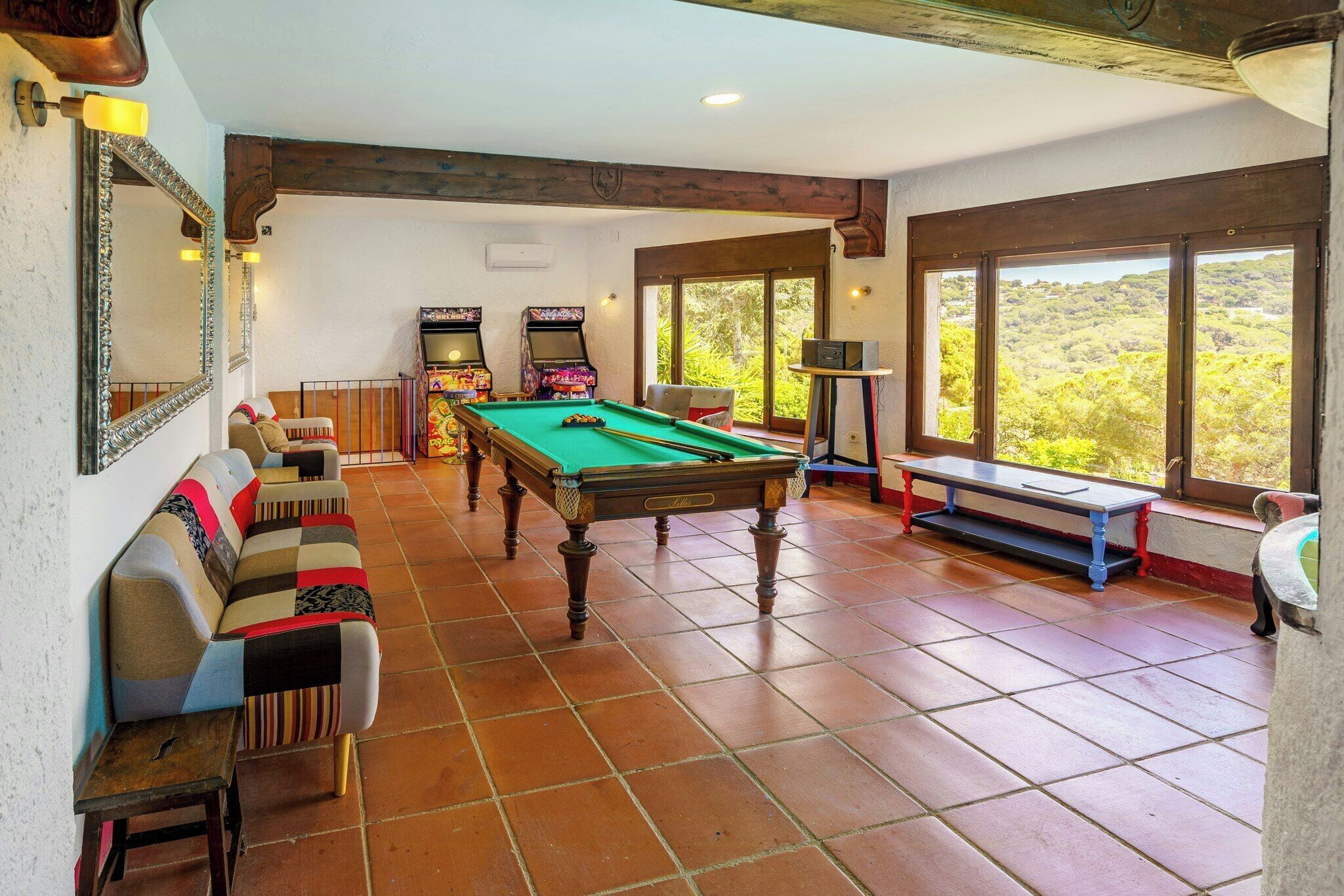A pool table in a room with a sofa, arcade games, and large windows.