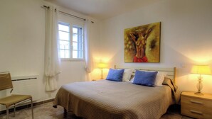 Un dormitori amb un llit, una cadira, una finestra amb cortines, un quadre a la paret i làmpades de nit.