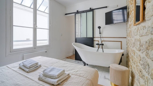A bedroom with a large bed, a bathtub, a TV, and a stone wall. - Loft in Sentier - Paris - 6P - 2BR (Paris)