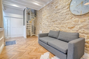 A living room with a stone wall, a grey sofa, a wooden herringbone floor, a staircase, and a large clock. - Loft in Sentier - Paris - 6P - 2BR (Paris)