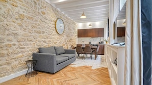 A living room with a stone wall, a grey sofa, wooden flooring, and a kitchen in the background. - Loft in Sentier - Paris - 6P - 2BR (Paris)