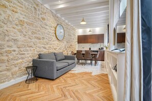 A living room with a stone wall, a grey sofa, wooden flooring, and a kitchen in the background. - Loft in Sentier - Paris - 6P - 2BR (Paris)