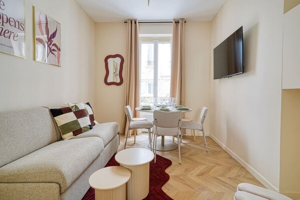Una sala de estar con un sofá, una mesa de comedor con sillas, un televisor montado en la pared y un ventanal con cortinas.