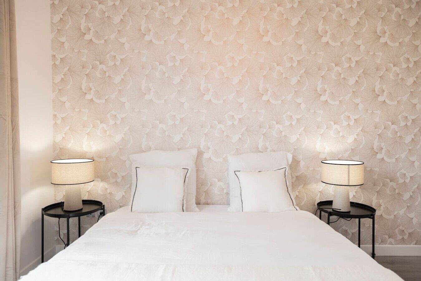 Quarto com papel de parede estampado, duas mesas de cabeceira com abajures e uma cama com roupa de cama branca.