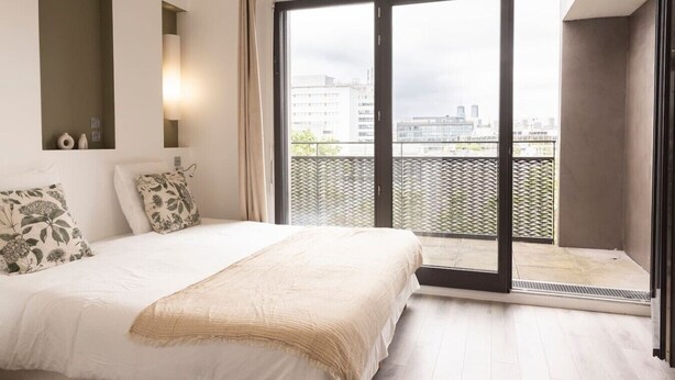 Un dormitorio con cama, ventana y vistas a edificios.