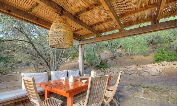 Un tavolo in legno all'aperto con sedie, un lampadario intrecciato e un muro in pietra sullo sfondo.