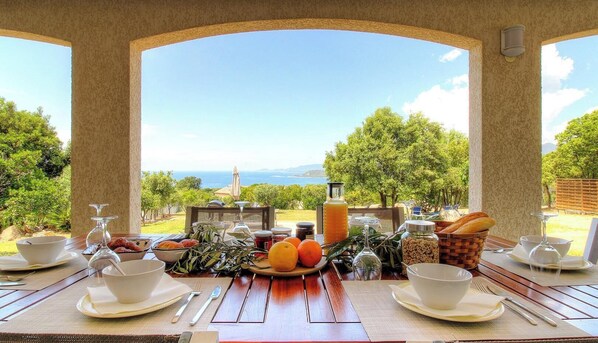 Ein Essbereich im Freien mit Blick auf das Meer, ein Tisch gedeckt mit Tellern, Gläsern und Früchten.