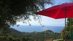 En terrasse med to liggestole og en rød parasol med udsigt over et kystlandskab med bakker og havet.