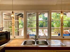 A kitchen with a double sink, a microwave, and a view of the outdoor deck through large windows. - The Pink Door - 3 Bedroom Cottage - Walking Distance to Town (Maleny)