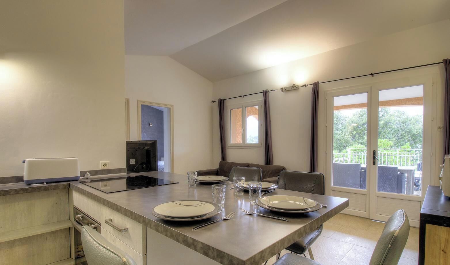 Una cucina moderna con isola centrale, zona pranzo con sedie e vista sul verde attraverso le finestre.