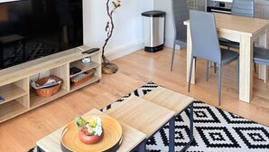 A modern living room with a flat-screen TV, a dining area with a wooden table and chairs, a kitchen with cabinets and a stove, and a decorative wall clock. - Beautiful setting with a terrace overlooking the sea (Saint-Cyprien)