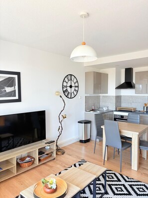 A modern living room with a flat-screen TV, a dining area with a wooden table and chairs, a kitchen with cabinets and a stove, and a decorative wall clock. - Beautiful setting with a terrace overlooking the sea (Saint-Cyprien)