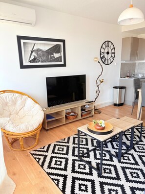 A living room with a television, a wicker chair, a coffee table, a patterned rug, and a kitchen area in the background. - Beautiful setting with a terrace overlooking the sea (Saint-Cyprien)