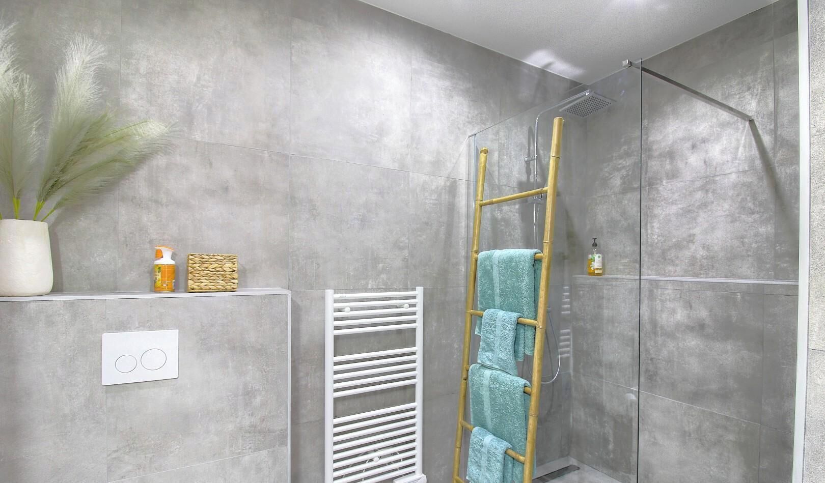 Ein modernes Badezimmer mit grauer Betonwand, einem weißen Handtuchhalter, einer Holzleiter und einem Duschbereich.