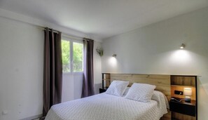 Ein Schlafzimmer mit Bett, Vorhängen, einem Fenster, einem Nachttisch mit Lampe und einer Wandleuchte.