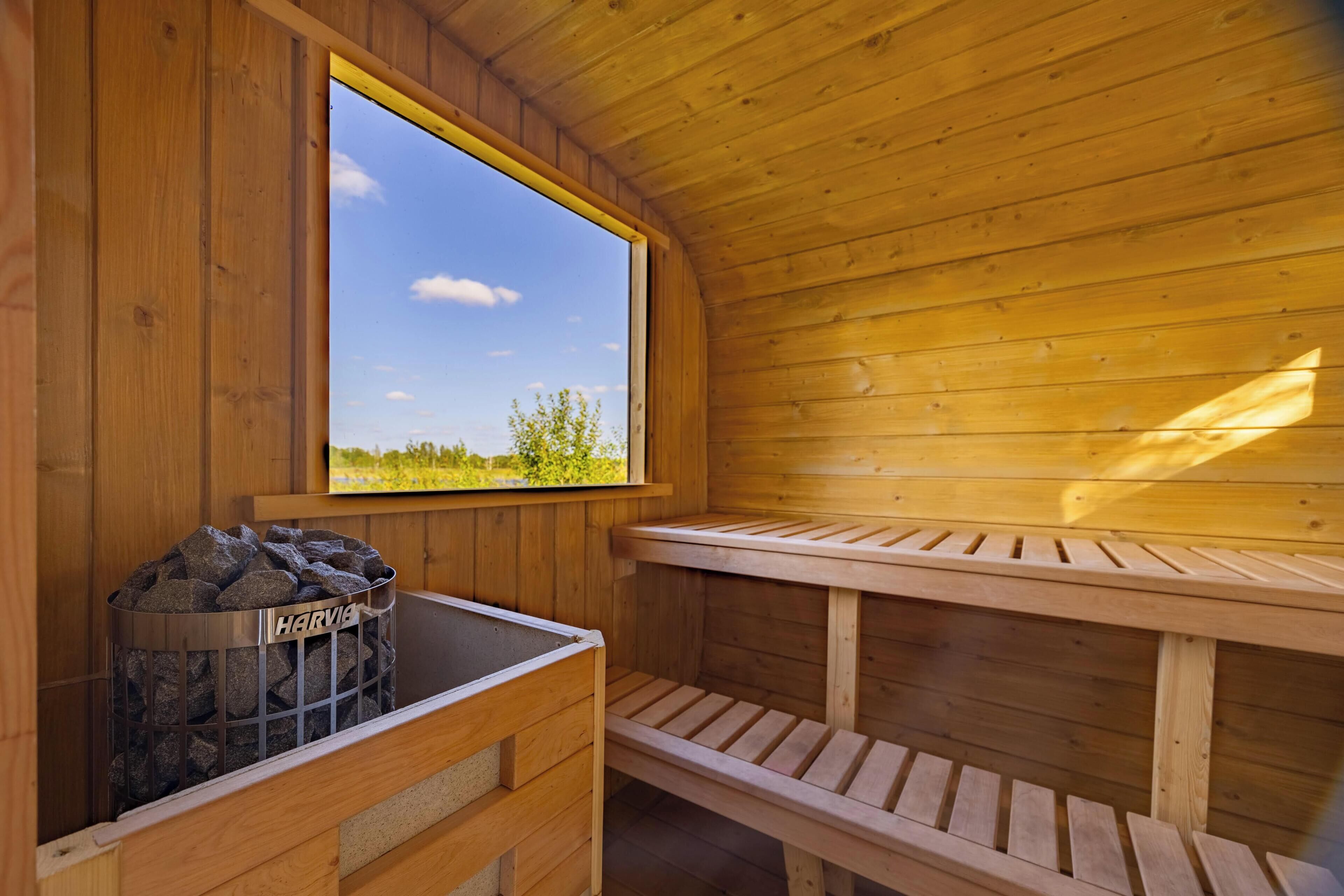 Una sauna de fusta amb una banca, una finestra que ofereix vistes a vegetació, i un contenidor metàl·lic amb l'etiqueta 'HARVIE'.
