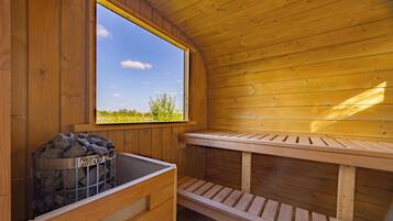 Una sauna de fusta amb una banca, una finestra que ofereix vistes a vegetació, i un contenidor metàl·lic amb l'etiqueta 'HARVIE'.