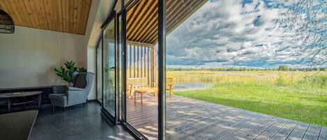 Un interior moderno con un amplio ventanal que da a una terraza exterior, techo de madera y vistas a un campo.