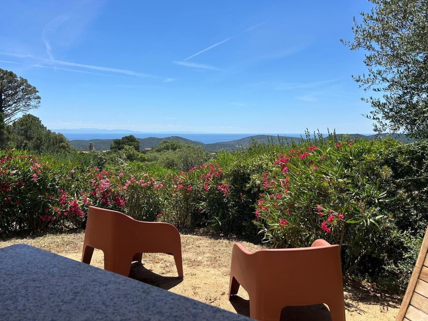 A terrace with two plastic chairs, a table, and a view of the sea and hills.
