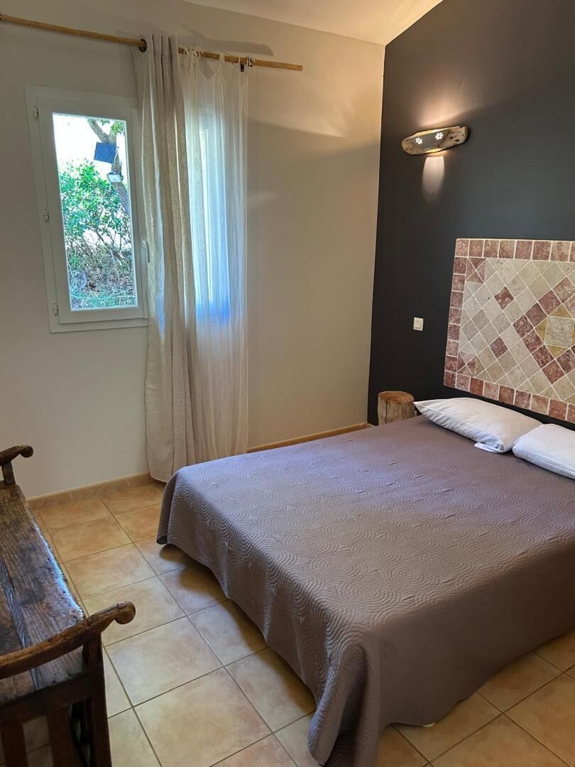 Un dormitorio con cama, ventana con cortinas, mesita de noche y lámpara de pared.