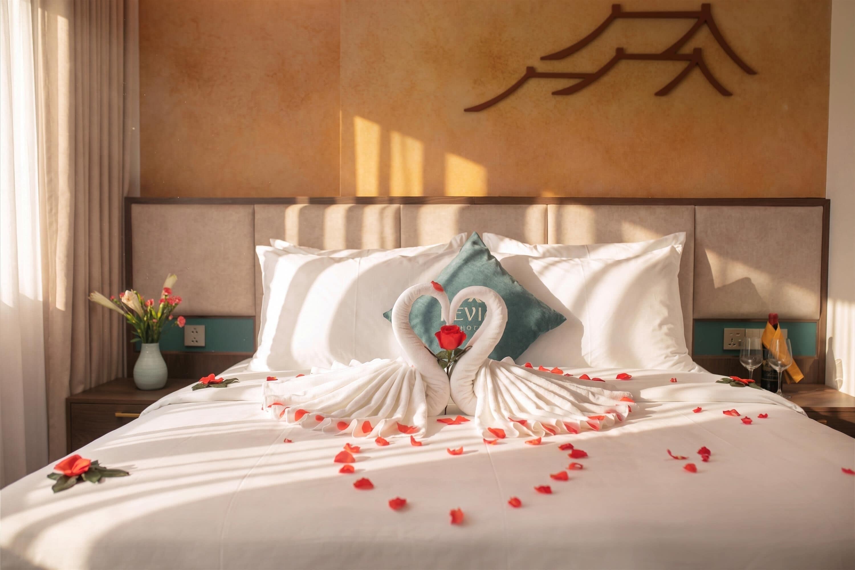 Un lit bien fait, orné d’un coussin en forme de cygne et de pétales de rose, un vase rempli de fleurs et un mur sur lequel est peint un grand caractère chinois.