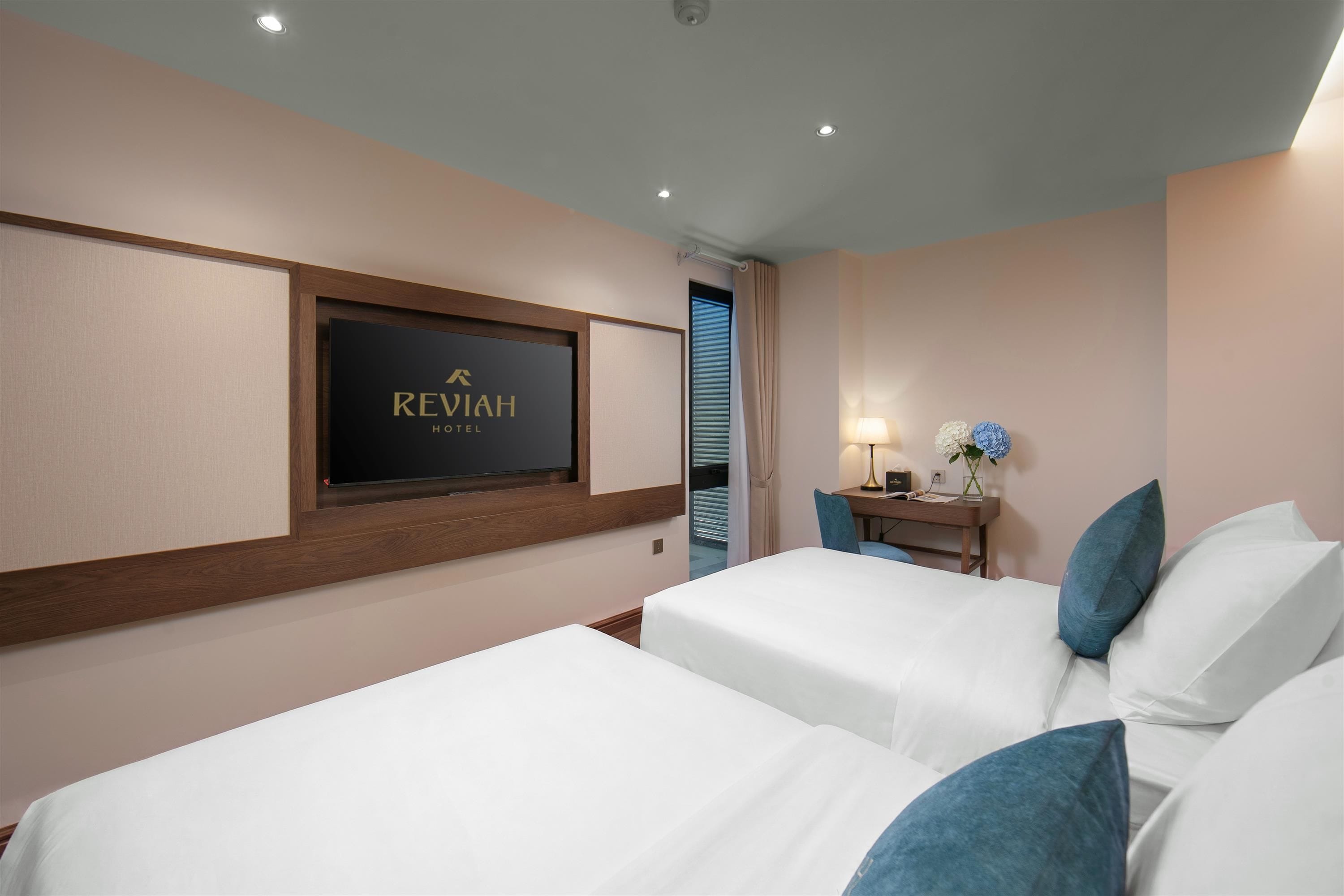 A hotel room with two beds, a TV mounted on the wall, a bedside table with a lamp, and a window with blinds.