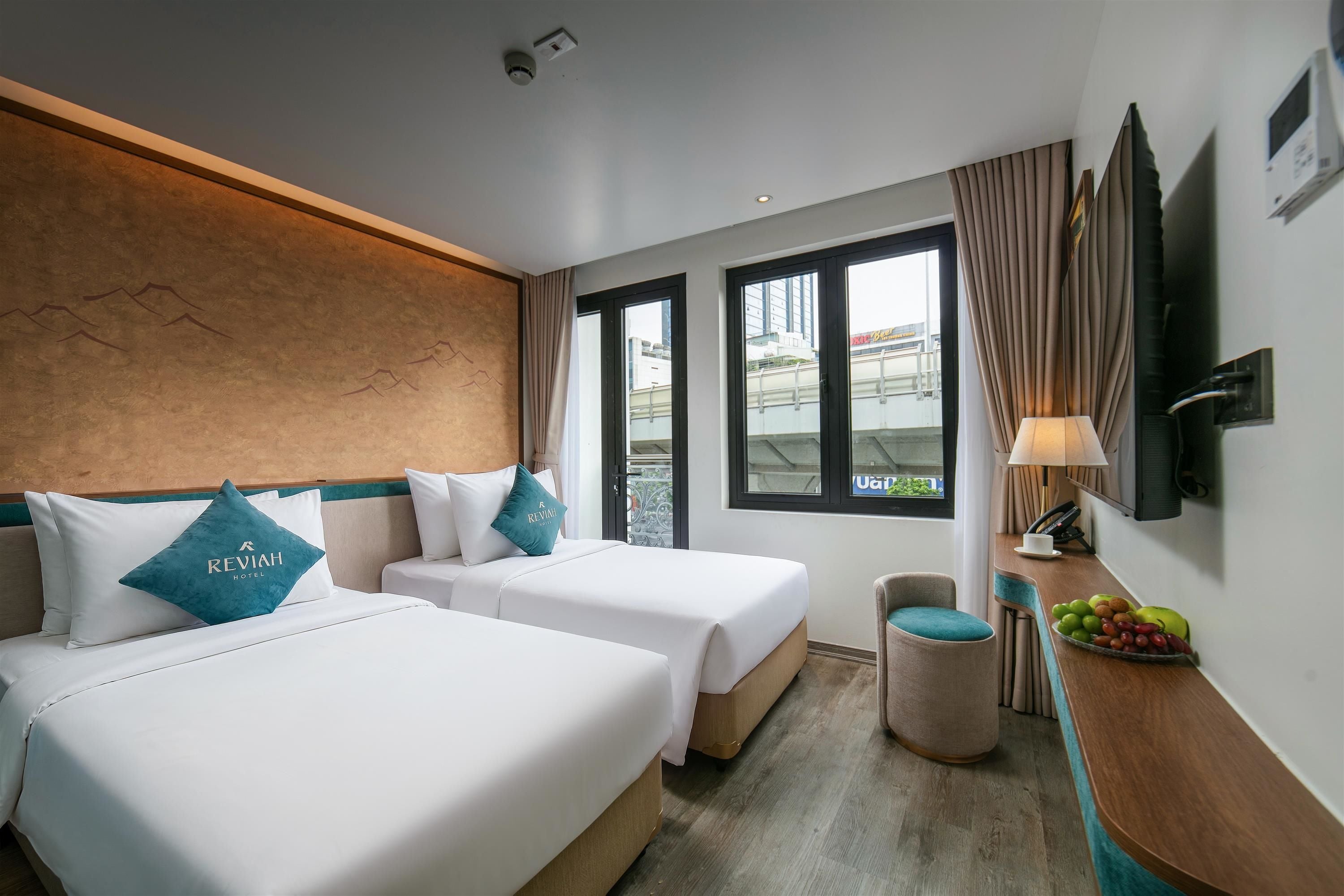 A hotel room with two beds, a desk with a lamp, a fruit bowl, and a view of the city through large windows.