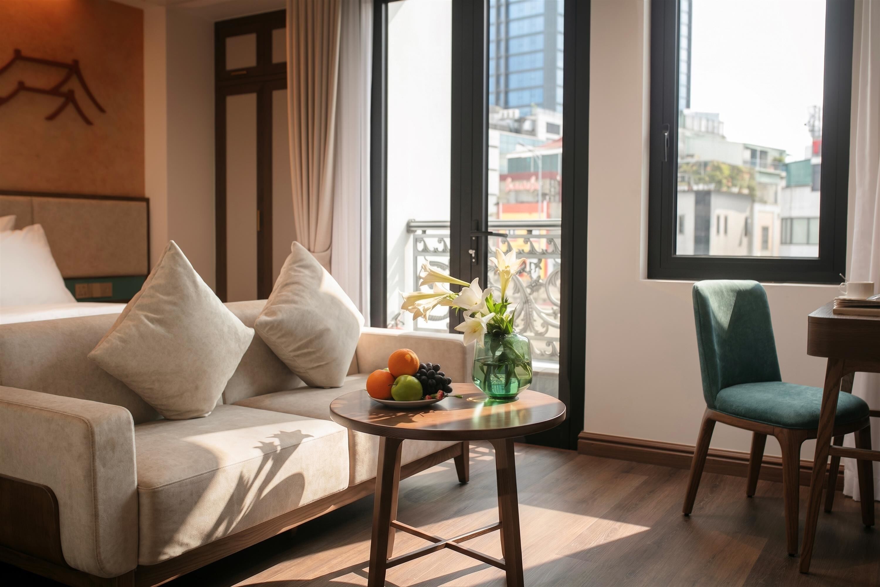 Une chambre d’hôtel moderne avec un canapé, une petite table ronde agrémentée de fruits et de fleurs, un fauteuil et une vue sur la ville grâce à de grandes fenêtres.