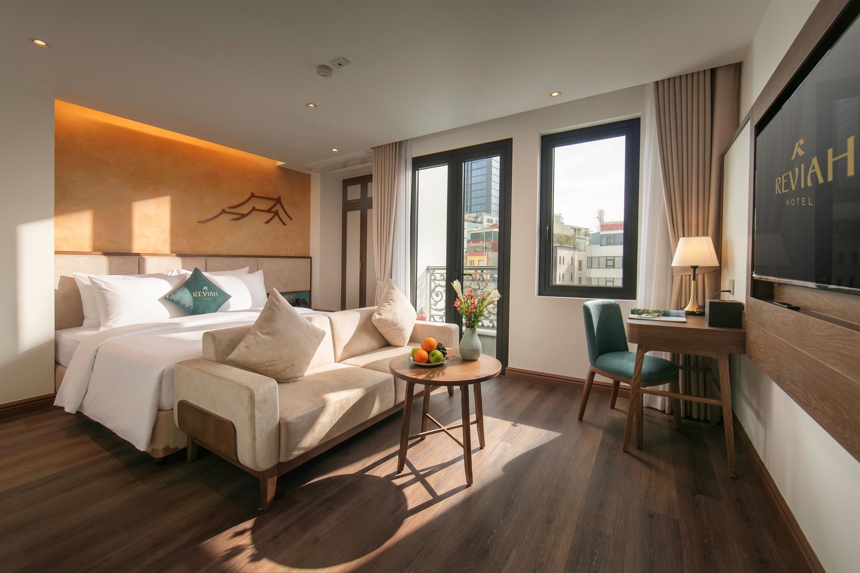 A hotel room with a bed, a sofa, a small table with fruit, a chair, and a TV on the wall.