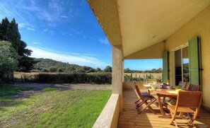 Eine überdachte Terrasse mit Tisch und Stühlen, mit Blick auf eine Rasenfläche mit Bäumen und einer Hügelkulisse.