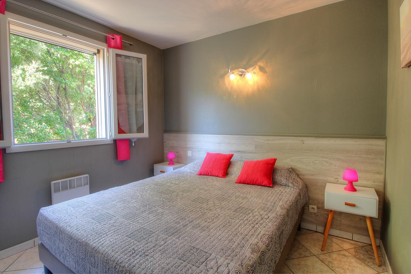 Une chambre à coucher avec un lit, une table de chevet, une lampe fixée au mur et une fenêtre avec des rideaux.