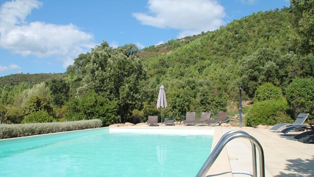 Una piscina con una escalera metálica, rodeada de sillas de descanso y sombrillas, con colinas verdes exuberantes al fondo.