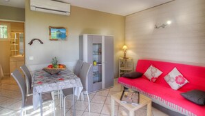 Un comedor con una mesa, sillas y una cesta de frutas. Hay un sofá rojo con cojines y una lámpara. Un equipo de aire acondicionado está montado en la pared.
