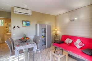 Een eetruimte met een tafel, stoelen en een fruitmand. Er is een rode bank met kussens en een lamp. Een airconditioner is aan de muur bevestigd.