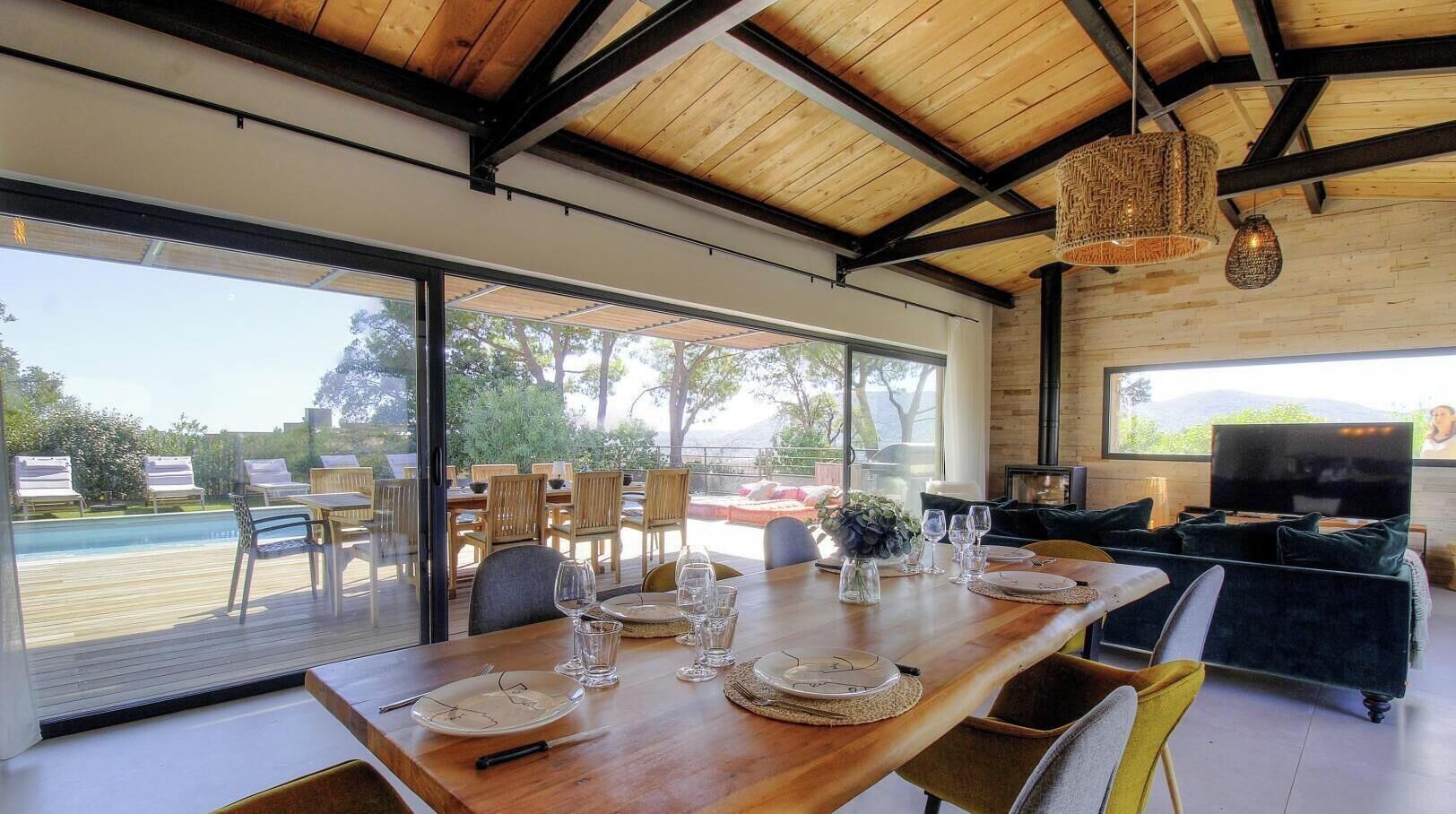 A dining area with a wooden table set for six, a sofa, and a TV in a modern interior with large windows.