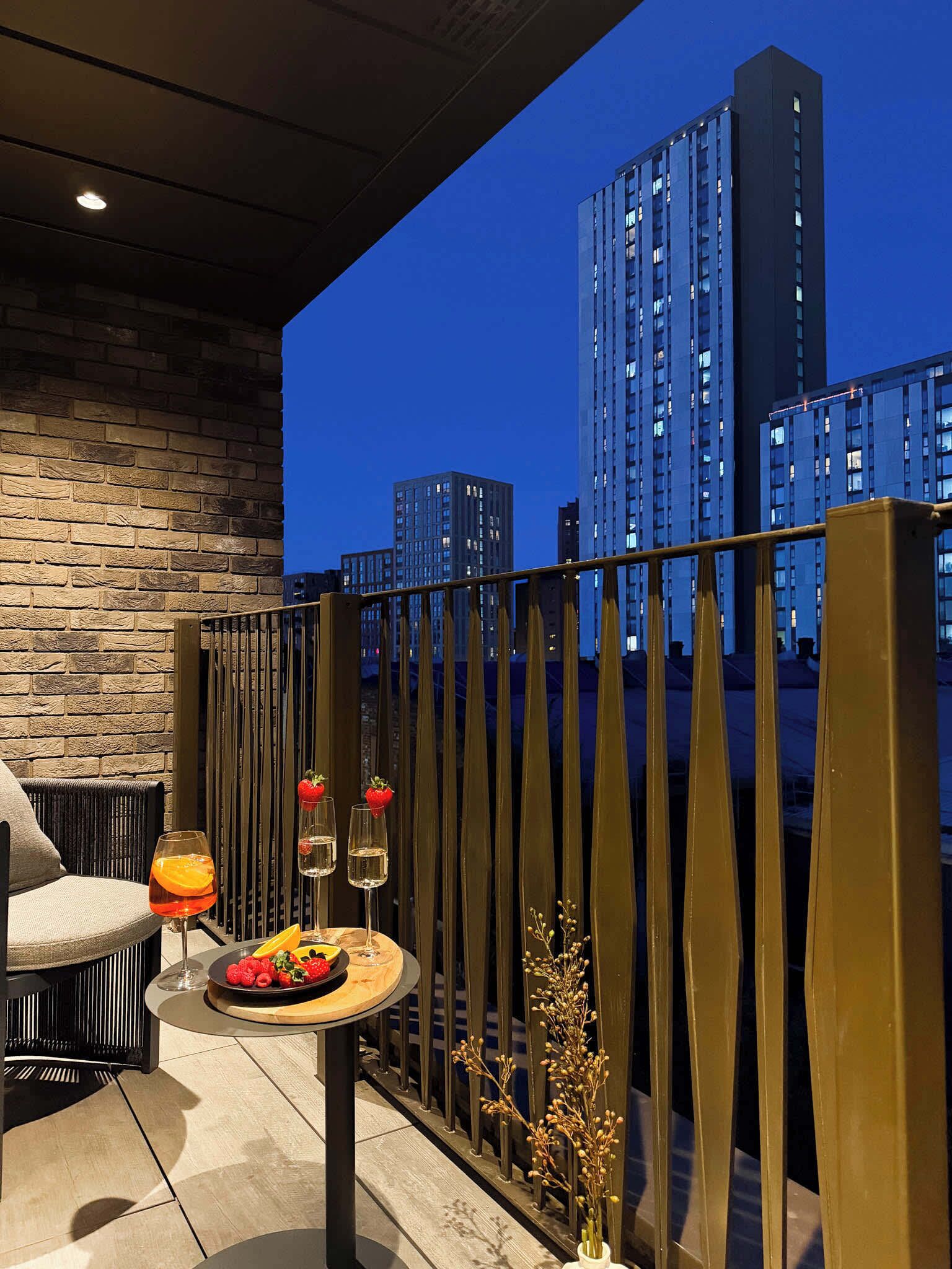 A balcony with a view of tall buildings at dusk, a table with a plate of strawberries and a glass of orange juice, and a vase with dried plants.