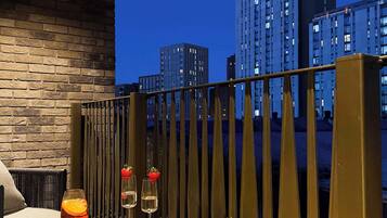 A balcony with a view of tall buildings at dusk, a table with a plate of strawberries and a glass of orange juice, and a vase with dried plants.
