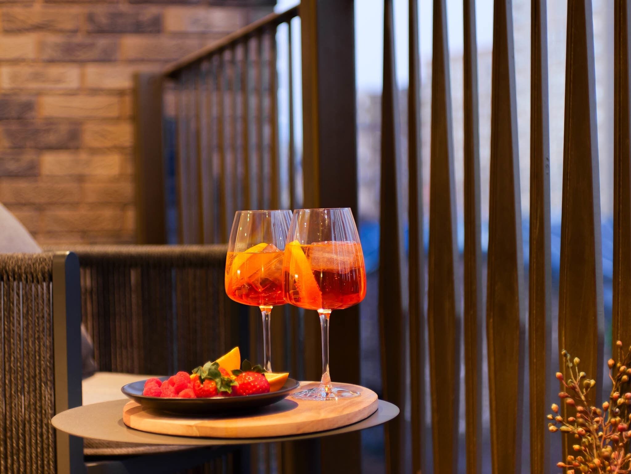 Two glasses of red beverage with fruit garnish on a wooden table with a plate of fruit and a vase with small plants.