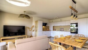 A modern living room with a flat-screen TV, a sofa, and a dining area with a wooden table set for four. - Le Figuier - FOLELLI (FOLELLI)