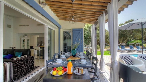 An outdoor dining area with a table set for six, surrounded by chairs, a wooden pergola, and a view of a swimming pool and garden. - Villa Loulou - BONIFACIO (BONIFACIO)