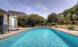 Ein rechteckiger Swimmingpool mit klarem, blauem Wasser, umgeben von einer Holzterrasse. Liegestühle und ein überdachter Bereich mit Tisch und Stühlen sind vorhanden.