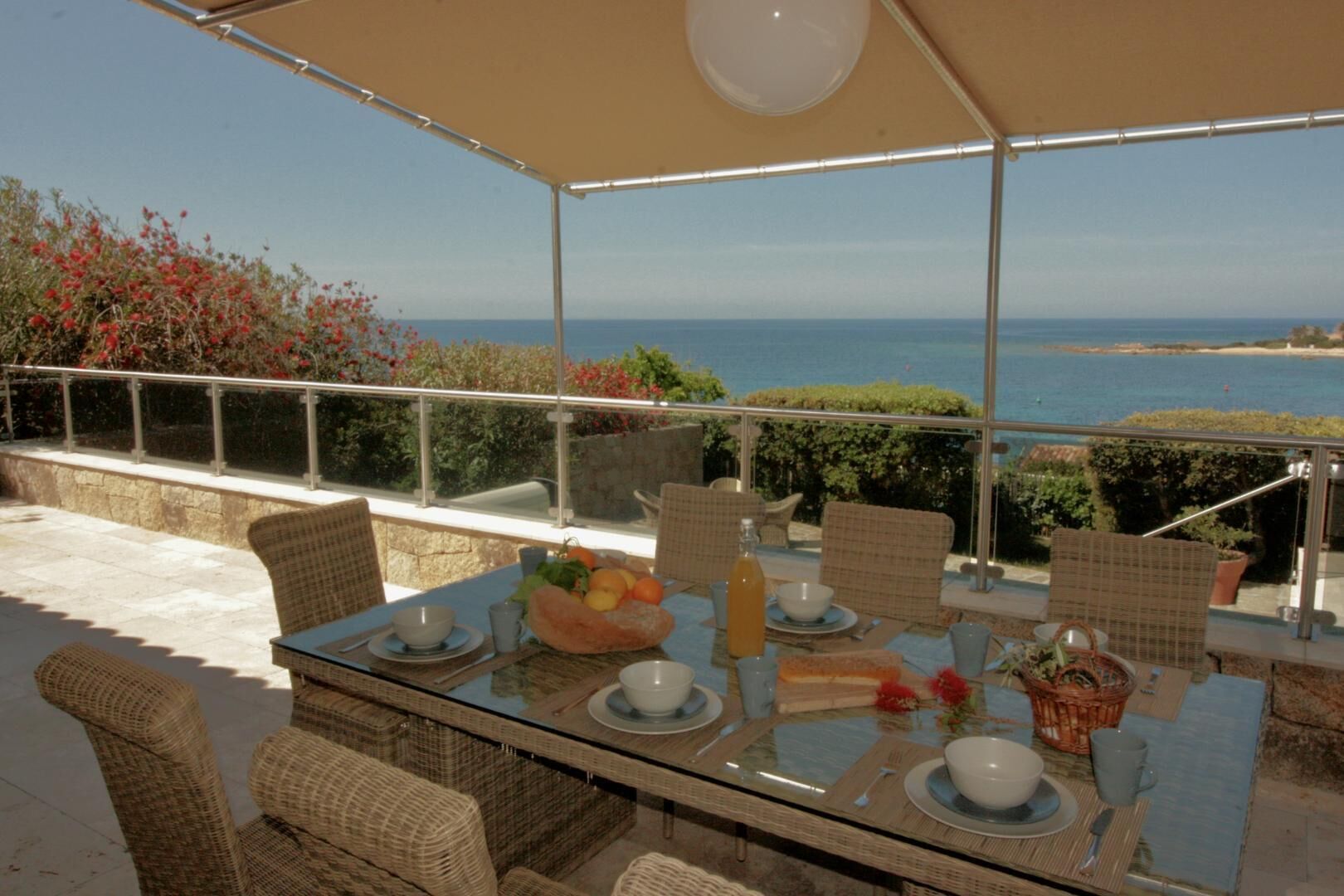 An outdoor dining area with a glass table, wicker chairs, and a view of the sea.