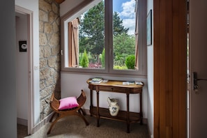 A room with a stone wall, a wooden chair with a pink cushion, a wooden table with books and a vase, and a view of a garden through a window.