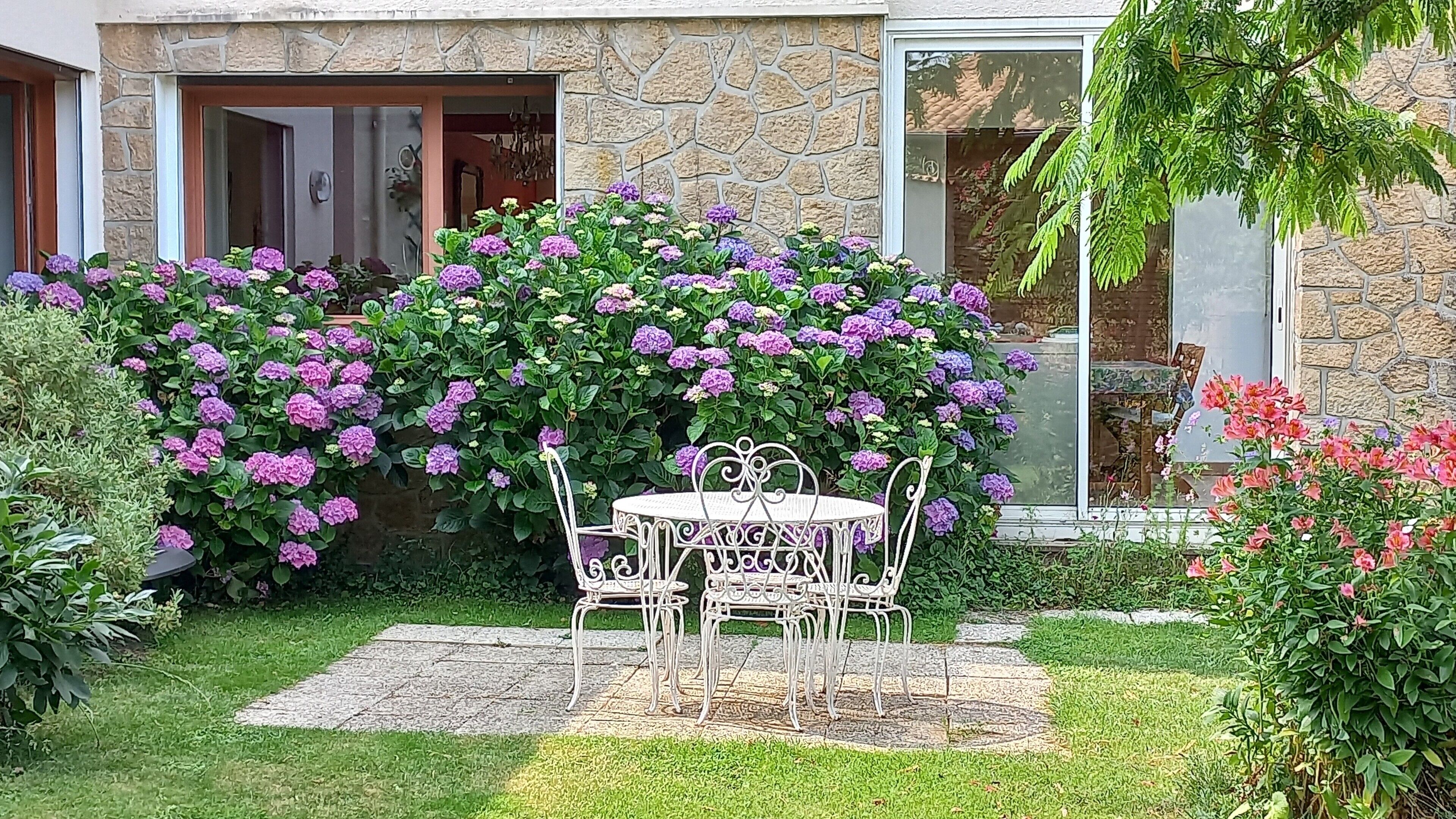 Une maison en pierre dotée d’un patio avec jardin, comprenant une table et des chaises en métal blanc, le tout entouré d’une végétation luxuriante et de fleurs violettes.