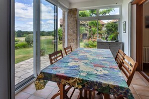 A dining area with a table covered in a floral tablecloth, surrounded by wooden chairs, and large windows offering a view of a garden and a house.