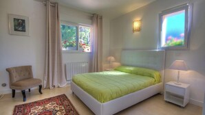Une chambre à coucher avec un lit, une chaise, une table de chevet, une lampe, une fenêtre et un radiateur.