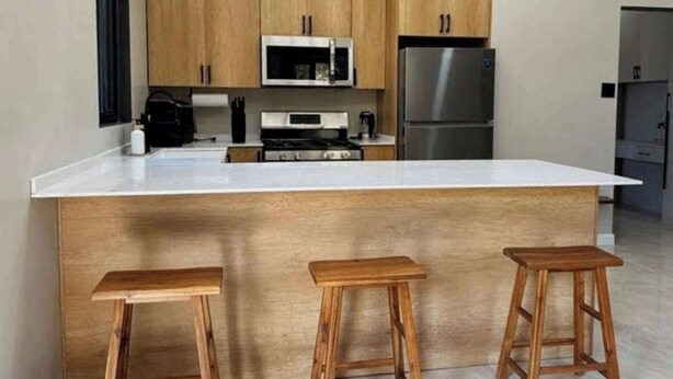 A kitchen with wooden cabinets, a white countertop, a stainless steel refrigerator, and three wooden stools. - Earth at Haven+ | Refined Island Escape (St.kitts)
