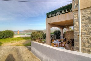 Ein Steinhaus mit überdachter Außenbestuhlung, Blick auf das Meer und einem entfernten Turm.